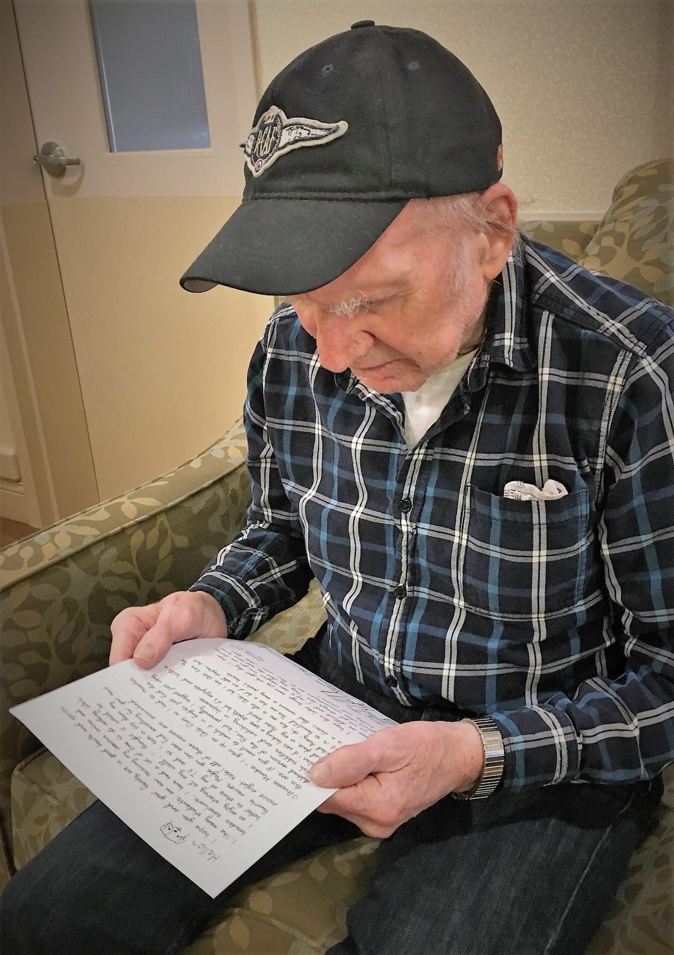 Old man wearing a baseball cap and plaid shirt sitting in a chair reading a letter
