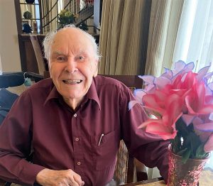 Senior man, smiling, balding, in a burgundy shirt, sitting next to a bouquet of pink flowers.
