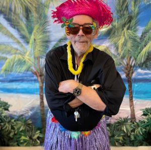 Senior man standing with arms crossed wearing a red hat, sunglasses, and a lei.