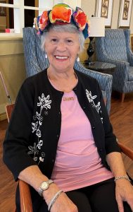 Older woman with short grey hair, sitting, wearing a pink t-shirt and black sweater with silver flower embroidery,