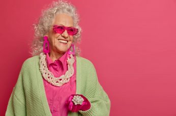 Charming fashionable senior woman in trendy sunglasses, jewelry looks aside with pleasant smile, has party for old people, celebrates something, poses