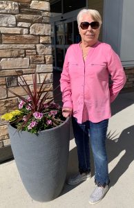 Senior woman standing next to a flower planter, wearing blue jeans, pink shirt, and sunglasses.