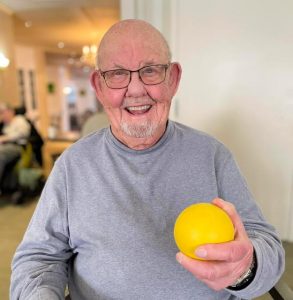 Older man in grey sweatshirt, wearing glasses smiling and holding a yellow ball.