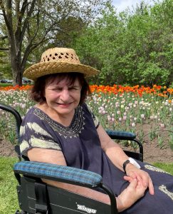Elderly woman with brown hair sitting in a wheelchair, wearing a straw hat, surrounded by flowers
