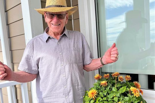 Senior man win straw hat, sunglasses, short sleeve shirt giving the thumbs up; balcony; summer container garden.