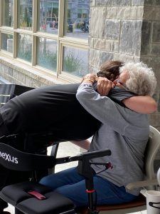 Caregiver in black bending down to hug older woman with dementia, siting with a walker