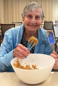 Older woman with short grey hair, wearing a blue cardigan, holding a whisk dipped in cookie dough, white mixing bowl with cookie dough