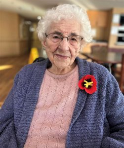 Older woman with short grey hair in a pink sweater and blue cardigan, wearing a poppy.