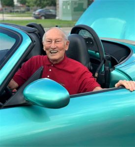 Senior man with white hair wearing a red golf shirt sitting in a blue sports care, laughing