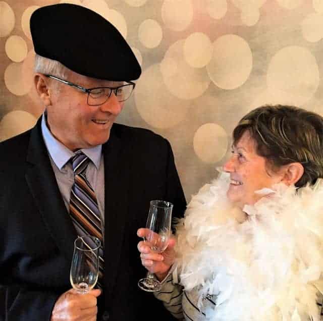Senior couple, man wearing black suit and hat, woman in white feather boa, looking at each other and holding champagne.