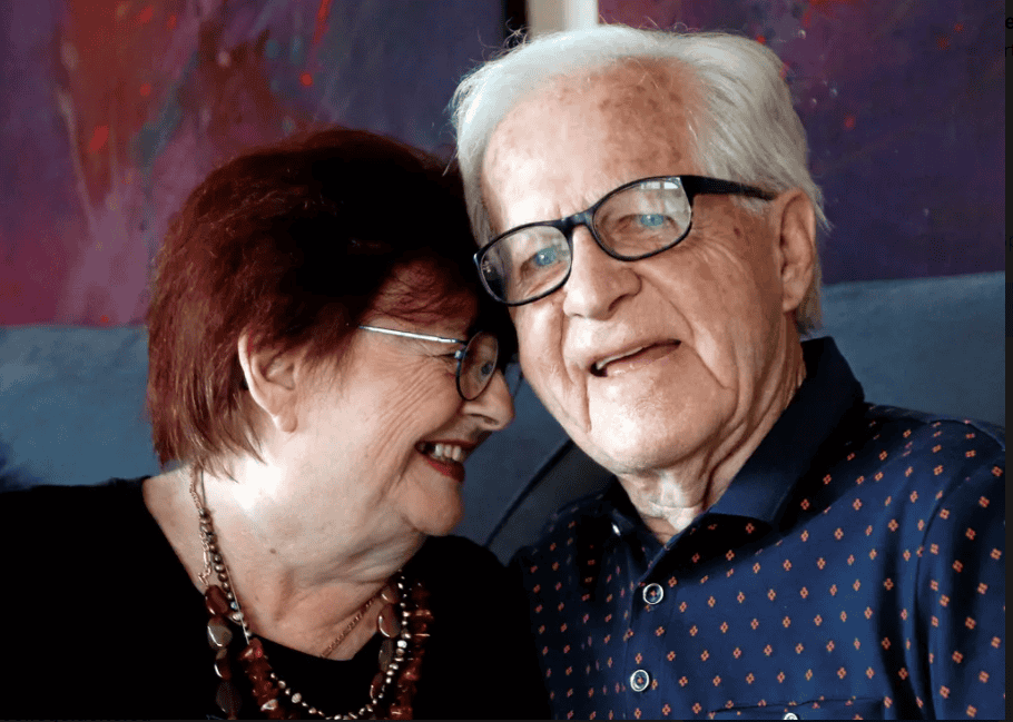 Love is in the Air - All Seniors Care Senior woman with red hair smiling while embracing a senior man with white hair and glasses.