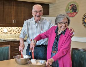 Senior couple cooking, man has arm around wife wearing pink cardigan.