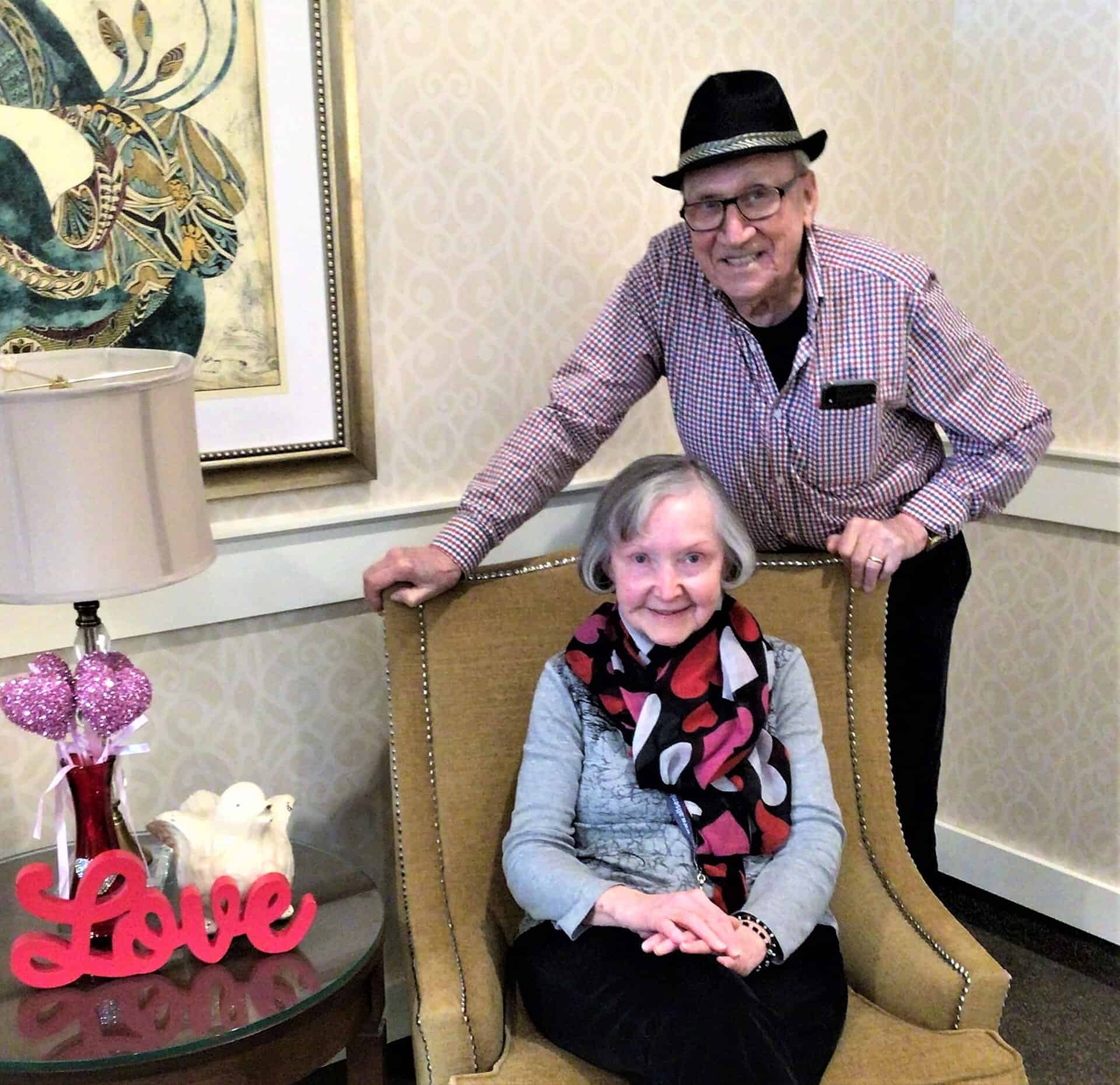 Love is in the Air - All Seniors Care Senior man in a black fedora stands behind a seated woman at a retirement residence.