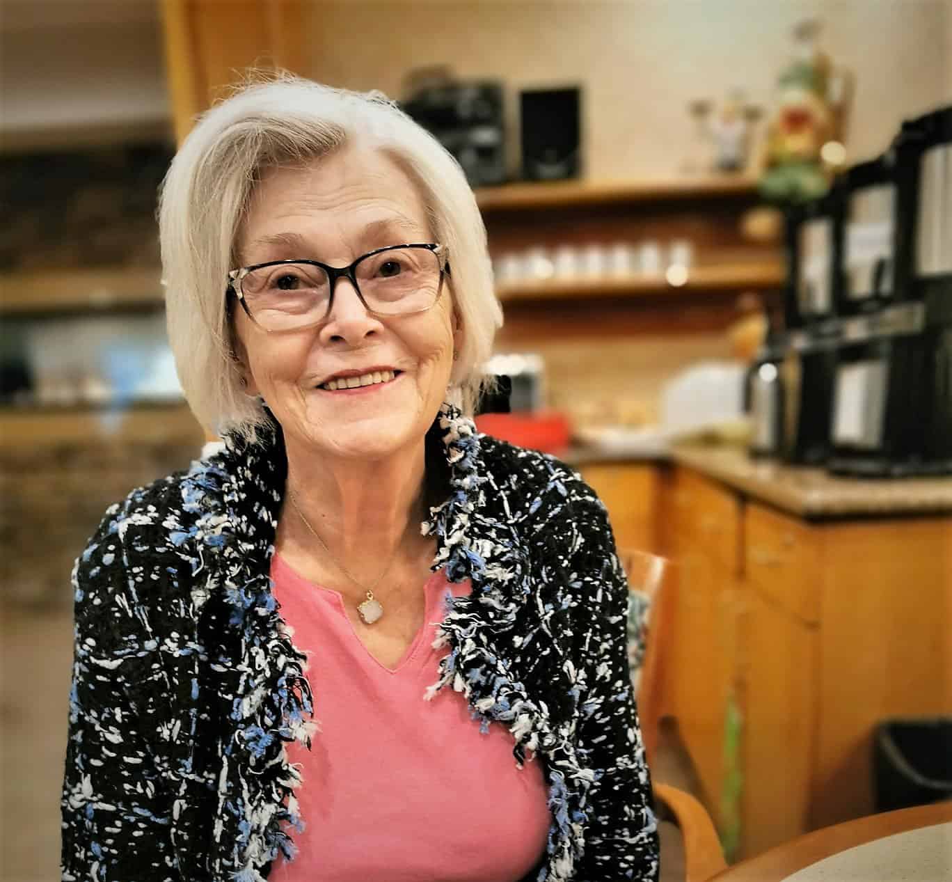 Woman with grey hair and glasses wearing a pink t-shirt and blue flowered blouse.