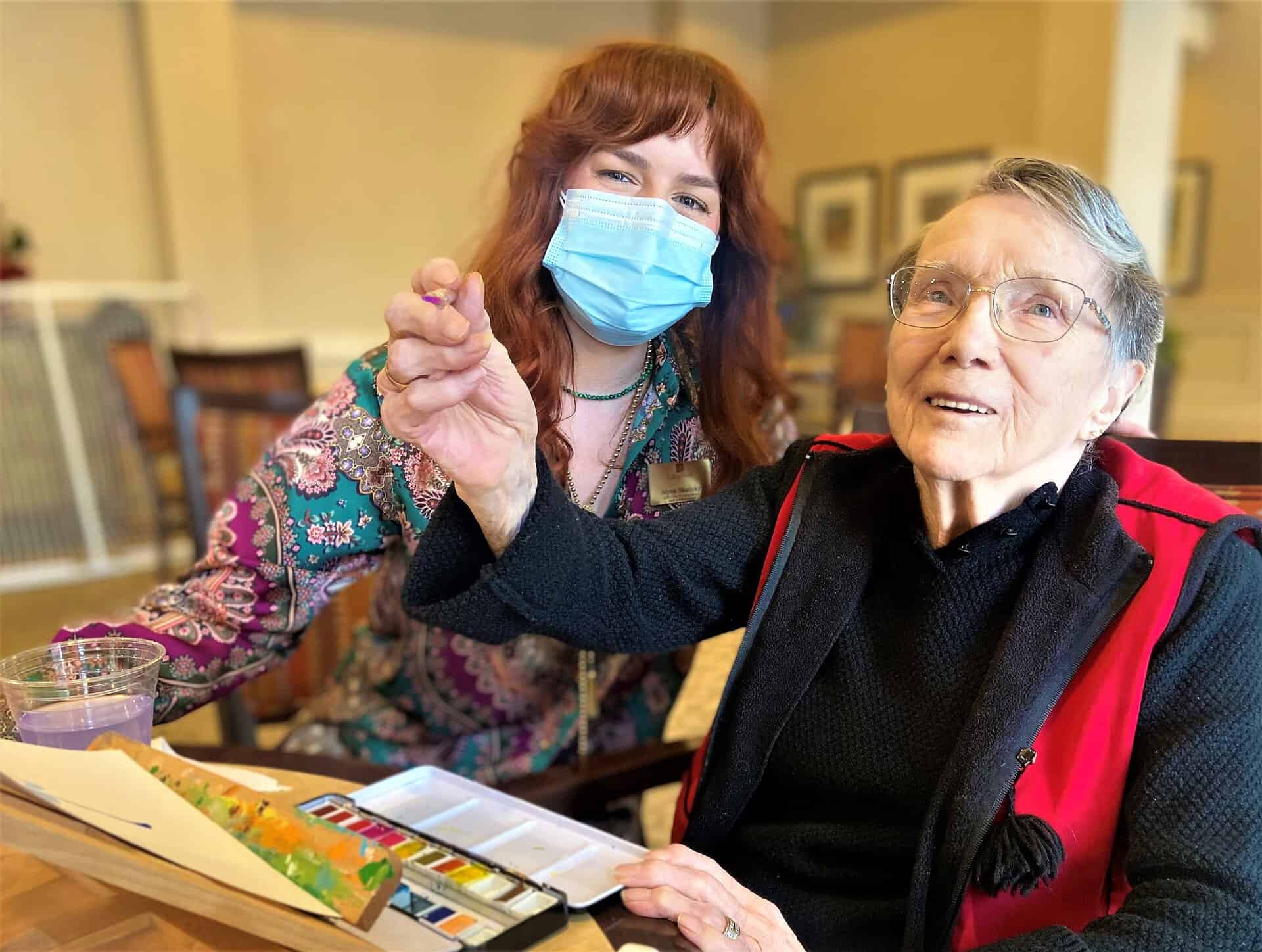 In The Spotlight: The Recreation Therapy A-Team - All Seniors Care Recreation therapist with long red hair next to a senior woman who is holding a paintbrush and doing a painting craft.